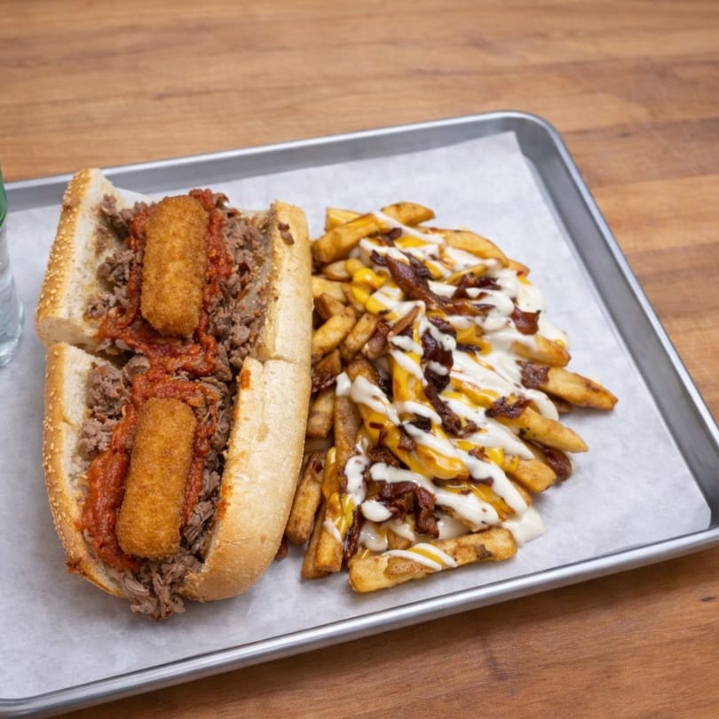 Cosmo's cheesesteak with mozzarella sticks and a side of loaded fries on a metal tray.