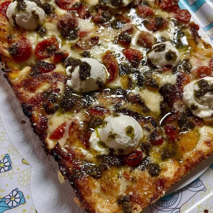 Close-up of a square pizza slice with pesto, cherry tomatoes, and fresh mozzarella on a patterned plate.